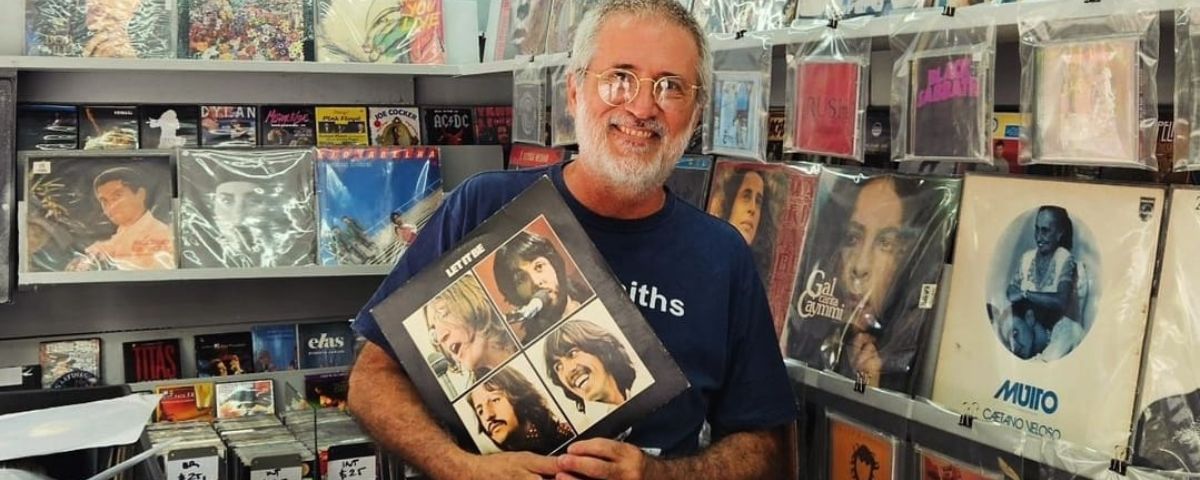 Homem sorrindo segurando disco de vinil dentro de uma loja de discos.