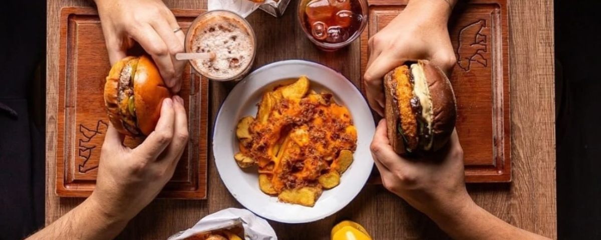 Mesa com duas pessoas segurando um hambúrguer cada uma, e também com batatas fritas e bebidas. 