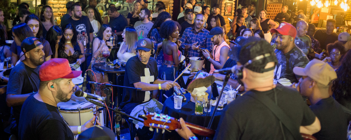 Banda tocando no bar Adega Original e público com diversas pessoas apreciando o som. 