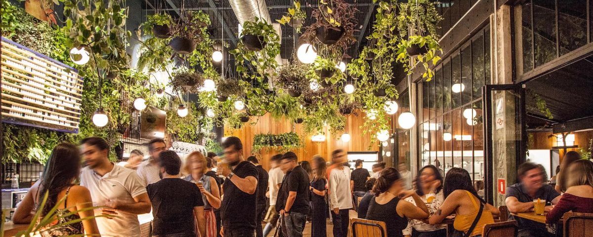 Diversas pessoas no Gaarden Bar, que possui plantas e luzes decorando o teto, além de ser um dos bares com música ao vivo. 