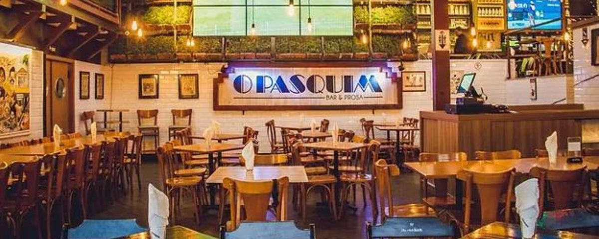 Mesas de madeira e diversas cadeiras no salão do Pasquim Bar e Prosa. 