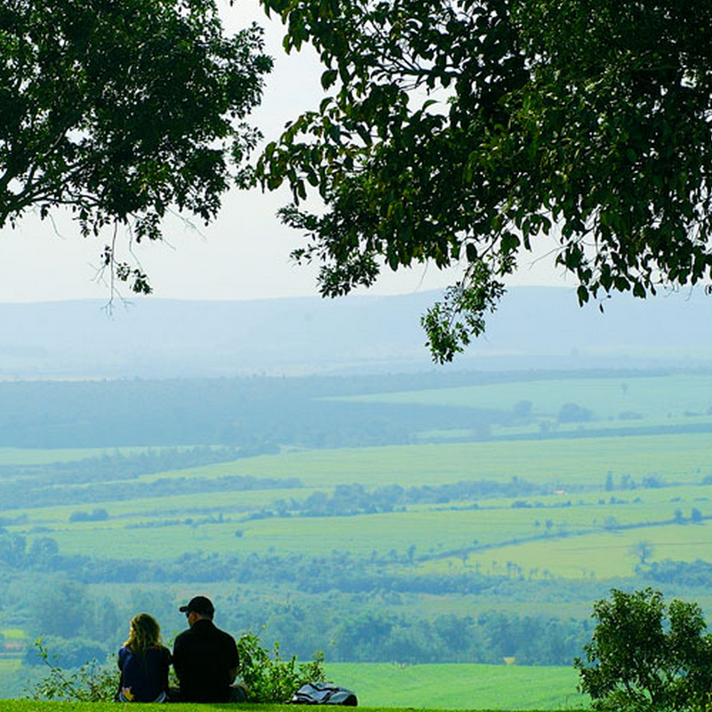 A imagem mostra um casal sentado no gramado contemplando as montanhas