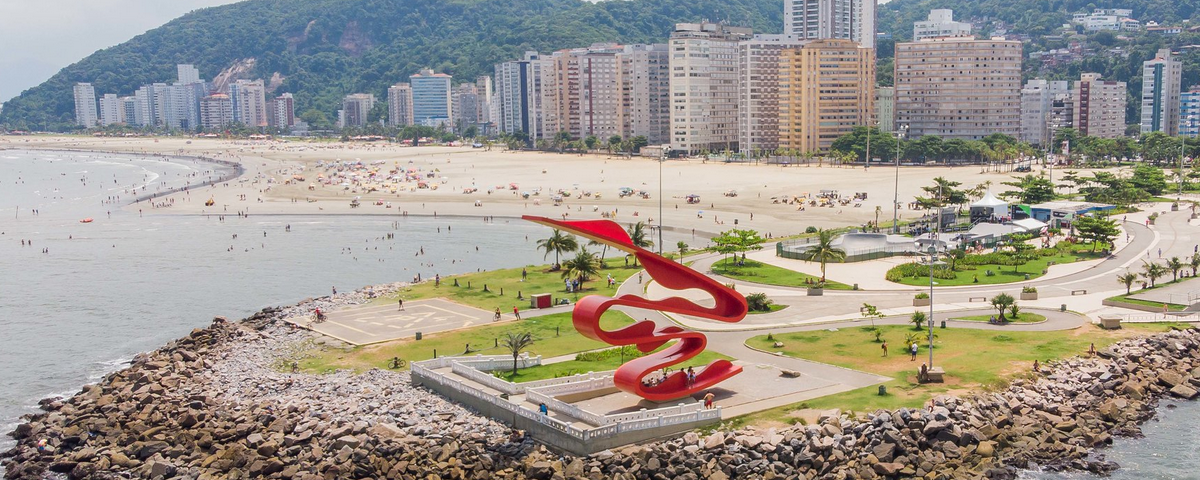 A imagem mostra a orla de uma praia, com prédios ao fundo e uma escultura vermelha 