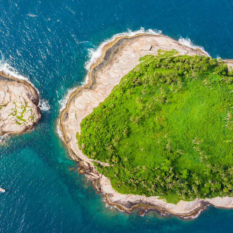 A imagem aérea mostra a costa da ilha com áreas verdes e o mar azul cristalino