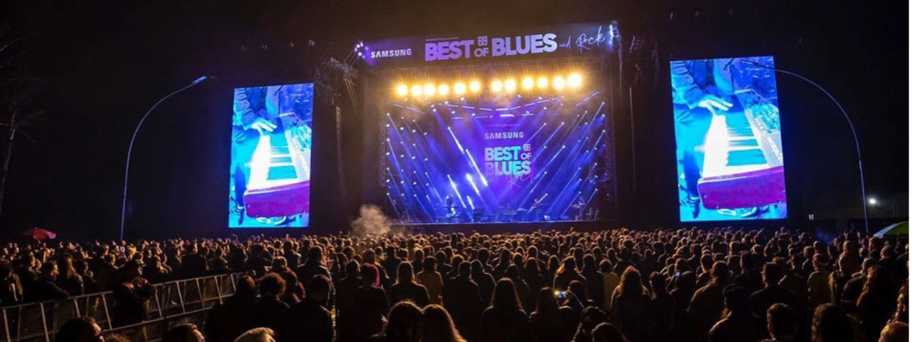 A imagem mostra a multidão no evento Best of Blues and Rock e ao fundo o palco