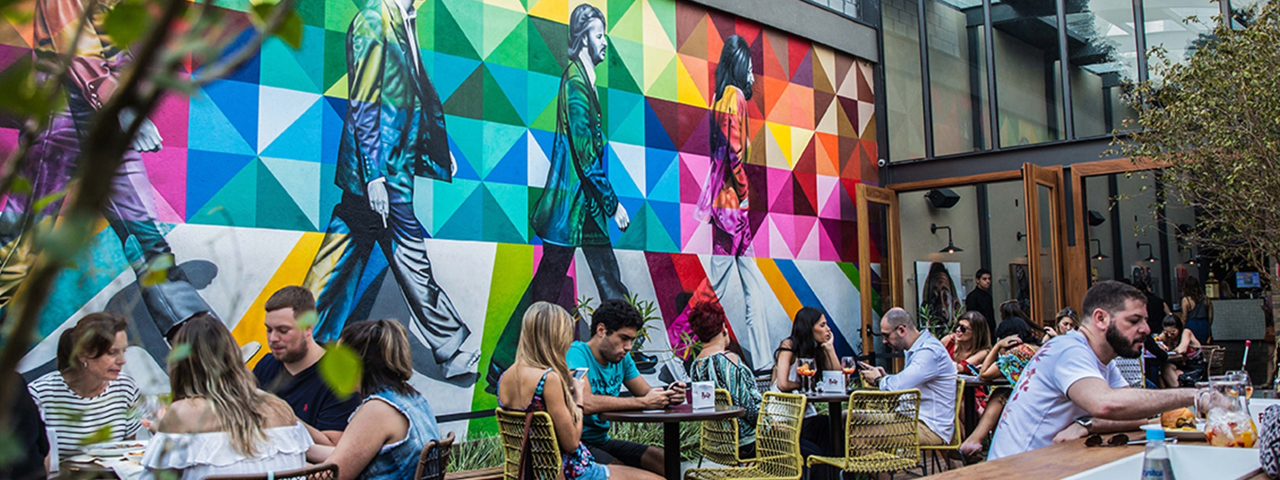 A imagem mostra o espaço externo do High Line Bar com pessoas sentadas na mesa e arte do Kobra ao fundo