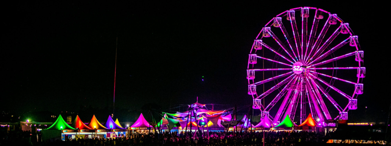 A imagem mostra o festival do Lollapalooza de noite e ao fundo as tendas com a roda gigante