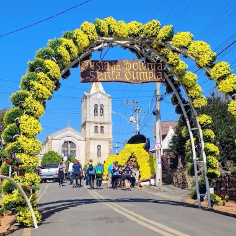 Portal de flores decorado da Festa da Polenta com a igreja da cidade ao fundo, celebrando a tradição no interior em julho