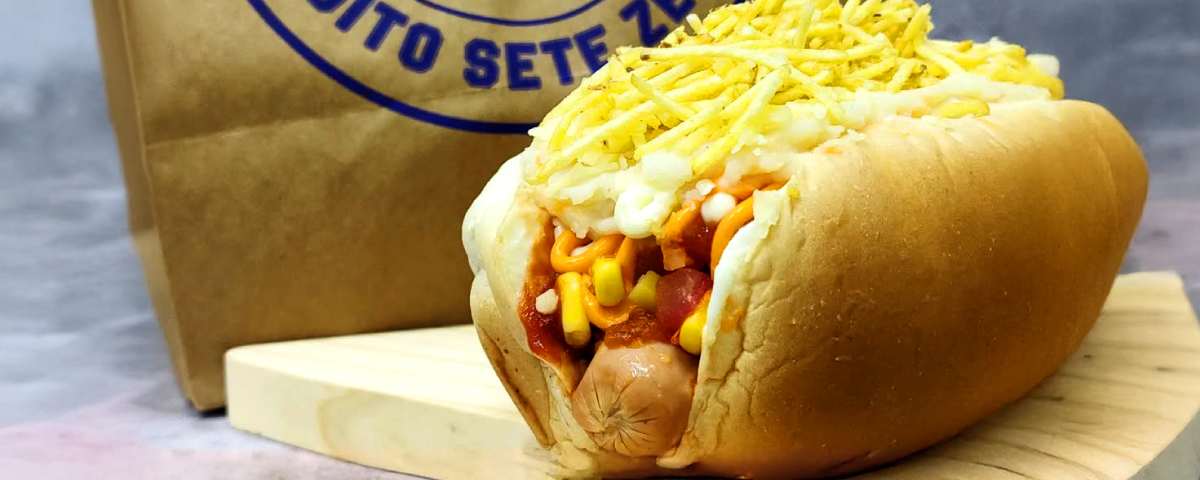 Hot dog coberto com batata palha crocante, em frente a uma sacola de embalagem personalizada, representando um estabelecimento com identidade visual marcante