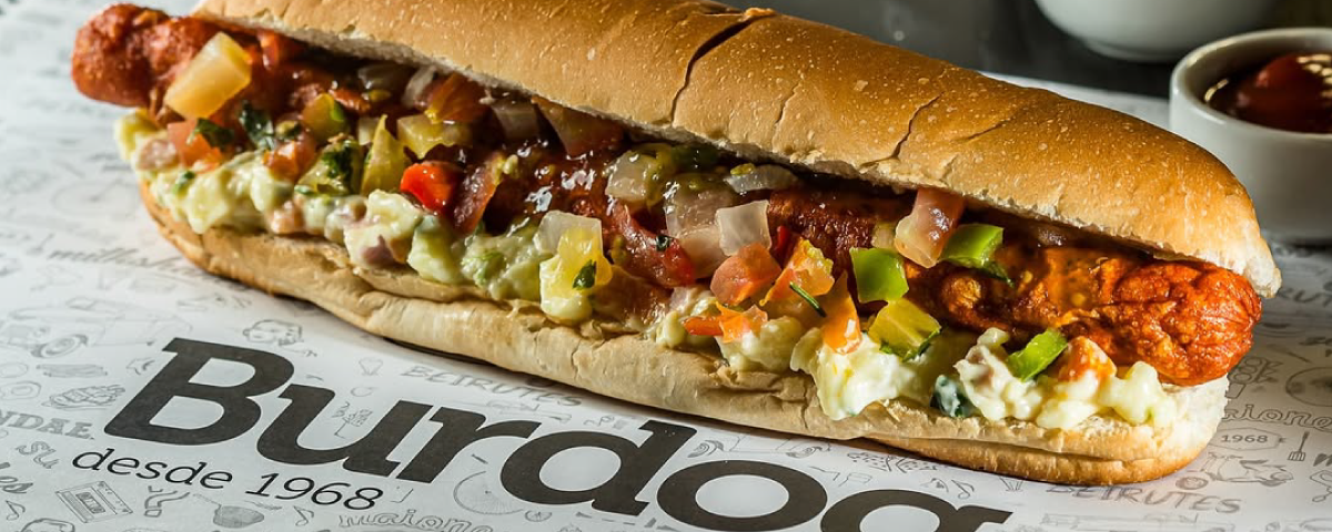 Hot dog do Burdog servido sobre uma mesa estampada típica da lanchonete, destacando a tradição e autenticidade do local