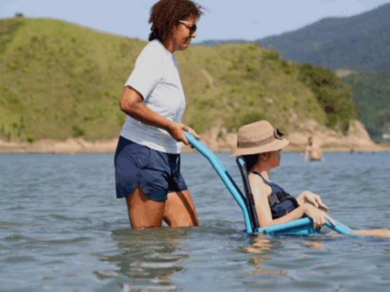imagem mostra uma mulher levando pessoa com mobilidade reduzida em cadeira especial em um passeio no mar