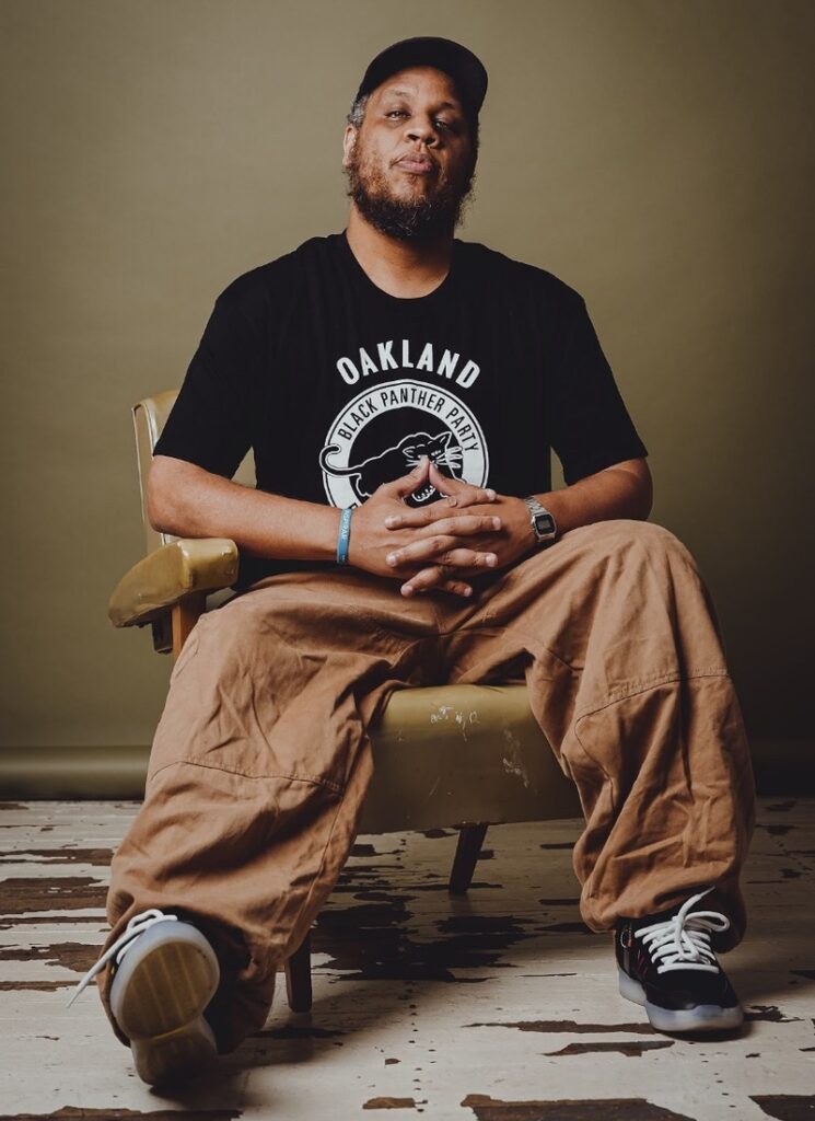 foto cantor Kamau sentado com blusa preta e calca bege