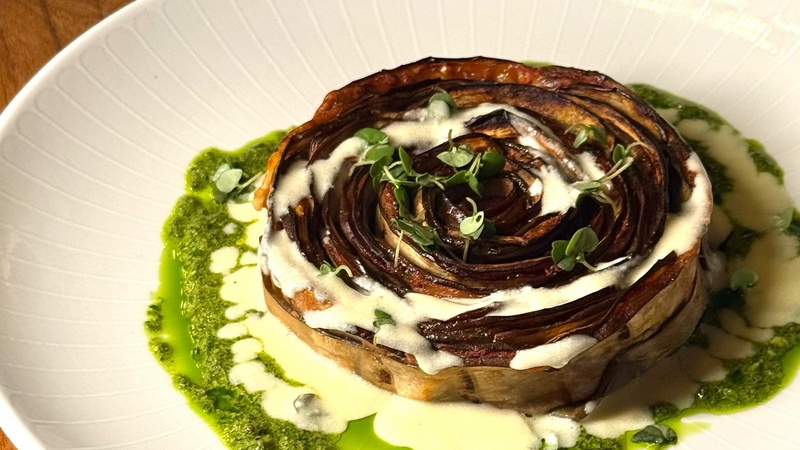 Prato de berinjela assada em formato de flor, com molho cremoso e azeite de ervas, finalizado com brotos verdes.