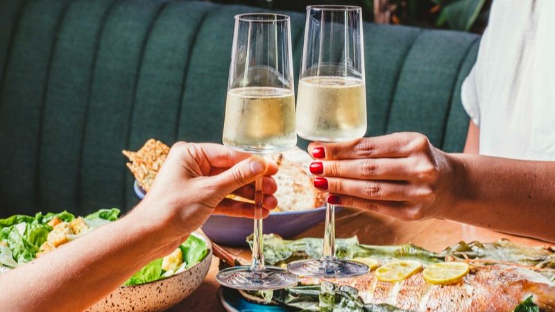 Duas pessoas brindam com taças de espumante durante almoço especial de Dia das Mães, em mesa com pratos e ambiente aconchegante de restaurante.