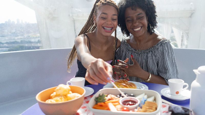 Mãe e filha participam de brunch dentro de cabine da Roda Rico, com mesa de café da manhã e vista da cidade de São Paulo ao fundo.