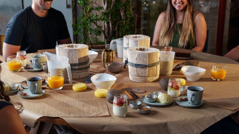 Participantes reunidos ao redor de uma mesa durante experiência gastronômica da Dengo Chocolates, com brunch servido em louças artesanais, sucos, frutas e peças de cerâmica em processo de criação.