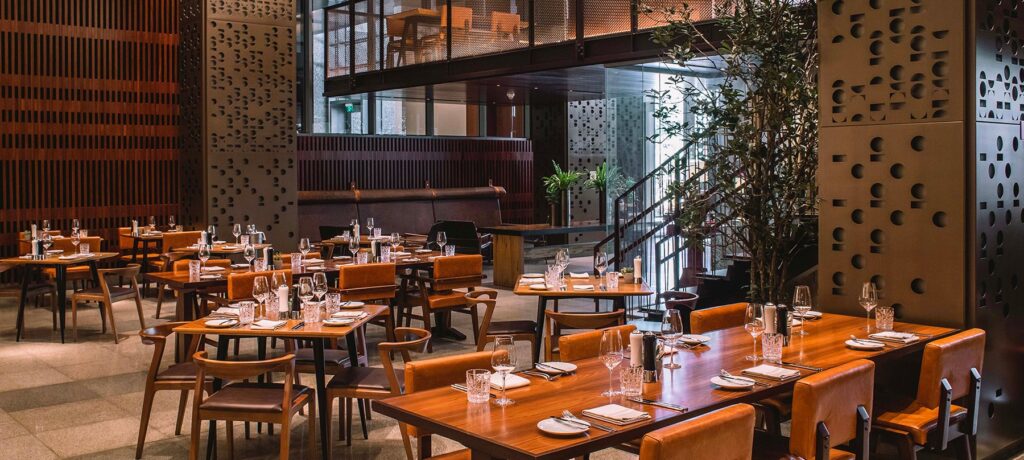 Salão interno do Restaurante Neto, com mesas de madeira postas, cadeiras em tons caramelo e ambiente moderno no JW Marriott Hotel São Paulo.