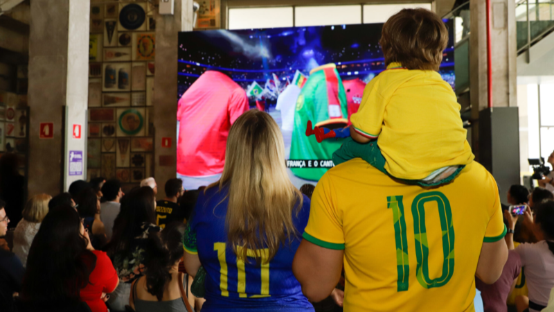 Família acompanha a transmissão de uma partida de futebol em um grande telão no Museu do Futebol. Em primeiro plano, duas pessoas vestem camisas da Seleção Brasileira, enquanto uma criança sentada nos ombros de um adulto observa a exibição. Ao redor, dezenas de visitantes assistem ao jogo em um ambiente interno decorado com elementos ligados à história do futebol.