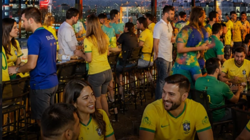 Foto de rooftop em São Paulo durante transmissão dos jogos da Seleção Brasileira na Copa do Mundo 2026. O espaço está decorado com bandeiras do Brasil e reúne dezenas de torcedores vestindo camisetas verde-amarelas. Pessoas conversam e acompanham a programação em mesas distribuídas pelo terraço, enquanto a paisagem urbana iluminada aparece ao fundo. A imagem ilustra opções de onde assistir à Copa do Mundo 2026 em São Paulo.