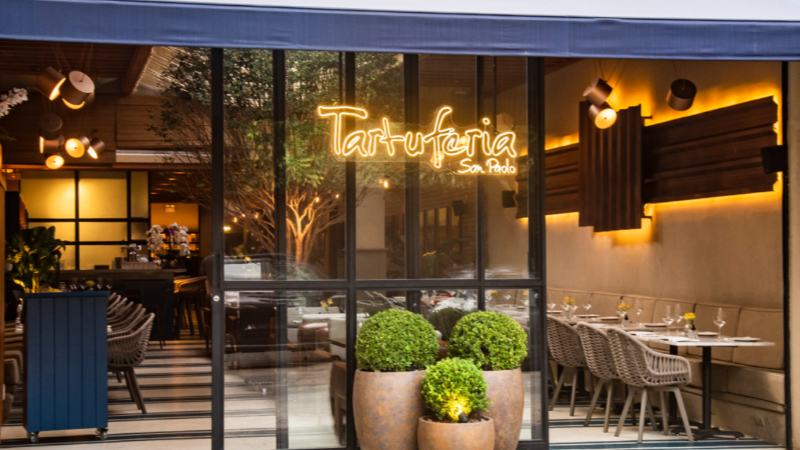 Fachada da Tartuferia San Paolo com amplas paredes de vidro, mesas do salão visíveis ao fundo, iluminação aconchegante e letreiro luminoso com o nome do restaurante.