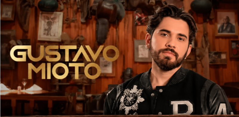 Imagem promocional do cantor Gustavo Mioto. O artista aparece em primeiro plano usando jaqueta preta, diante de um ambiente com decoração inspirada no universo sertanejo, com paredes de madeira e objetos rústicos. À esquerda está escrito o nome do cantor em letras douradas.