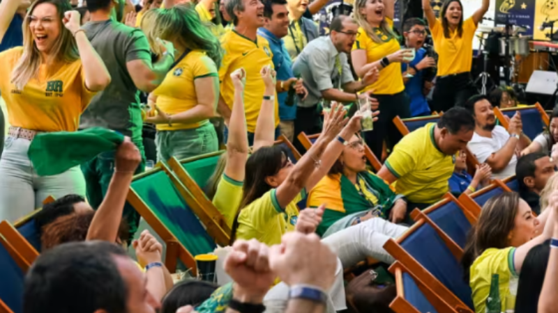 Torcedores vestidos com camisetas amarelas da seleção brasileira comemoram um lance durante a transmissão de uma partida de futebol. As pessoas estão sentadas em cadeiras de praia e espalhadas por um espaço aberto, com braços erguidos, sorrisos e expressões de entusiasmo, criando um ambiente de festa e confraternização.