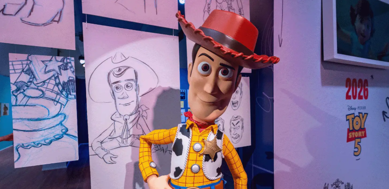 Personagem Woody, da franquia Toy Story, em tamanho ampliado, posa à frente de painéis com esboços originais do personagem. Ao fundo, há ilustrações do processo criativo da animação e um painel com a identidade visual de Toy Story 5, compondo um dos ambientes da exposição.