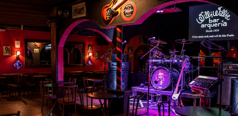 Interior do Willi Willie Bar e Arqueria, com mesas distribuídas pelo salão e palco equipado com bateria, guitarras, amplificadores e microfones preparados para apresentações ao vivo. O ambiente é iluminado por luzes em tons de vermelho e roxo, com decoração inspirada na cultura do rock.