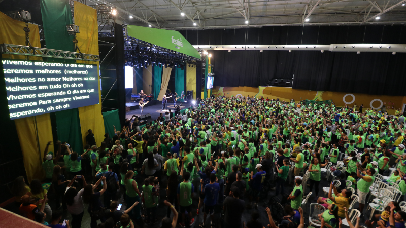 Público acompanha apresentação musical em grande espaço coberto durante a Festiva – O Nosso Mundial. Centenas de pessoas vestidas com camisetas verdes assistem ao show em frente ao palco, decorado com elementos visuais inspirados na Copa do Mundo.