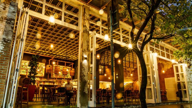 Fachada do Tuy Bar, Cocina com amplas portas abertas, iluminação em tons quentes, mesas internas e área de bar visível ao fundo durante a noite.