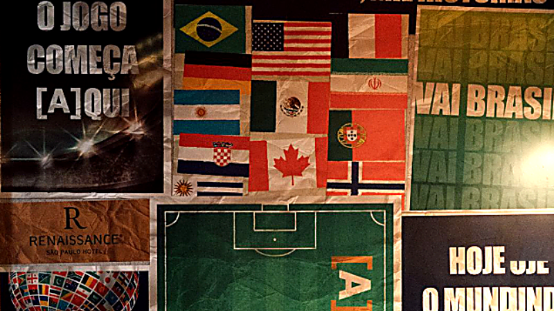 Painel decorativo da Arena 26, espaço temático do Renaissance São Paulo Hotel para transmissão dos jogos da Copa do Mundo 2026. A composição reúne bandeiras de diferentes países, frases relacionadas ao futebol e à união entre nações, além de uma representação de campo de futebol ao centro da imagem. O ambiente utiliza elementos visuais em tons de verde, azul e dourado inspirados no universo esportivo.