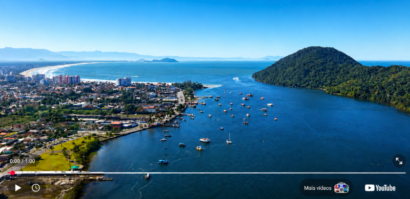 Vista aérea da Baixada Santista em um dia de céu limpo. Em primeiro plano, um canal de águas calmas com diversas embarcações ancoradas. À direita, um morro coberto por vegetação densa avança sobre o mar. À esquerda, a área urbana se estende até uma longa faixa de praia e o oceano. Ao fundo, montanhas formam o horizonte, compondo uma paisagem que integra natureza, cidade e litoral.