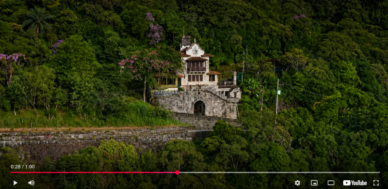 Vista aérea do Parque Caminhos do Mar, em Cubatão. Em meio à densa Mata Atlântica, uma construção histórica de pedra se destaca na encosta da serra, cercada por árvores de diferentes tons de verde. A imagem evidencia a integração entre patrimônio histórico e natureza preservada.