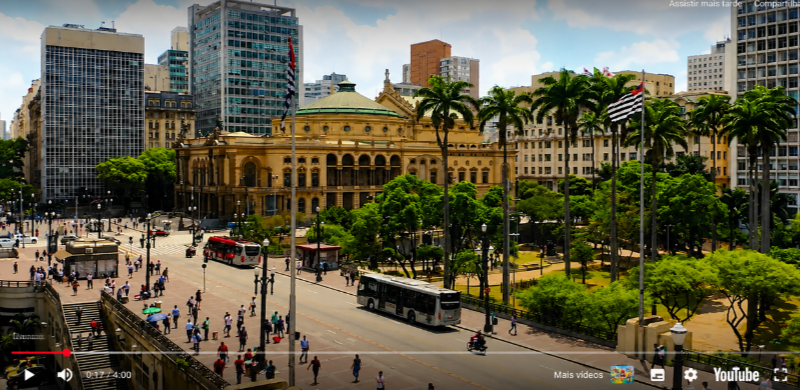 Vista ampla da Praça Ramos de Azevedo, no Centro de São Paulo, com o Theatro Municipal em destaque. O edifício histórico, de arquitetura eclética e cúpula esverdeada, é cercado por palmeiras, áreas verdes, ônibus, pedestres e edifícios altos, retratando o encontro entre o patrimônio cultural e a dinâmica da capital paulista.