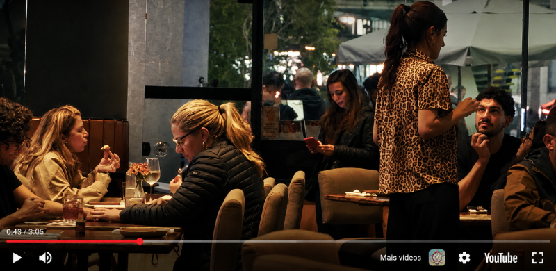 Ambiente interno do Bar da Onça com mesas ocupadas por clientes durante o período da noite. Pessoas conversam e fazem refeições enquanto uma atendente circula entre as mesas. Ao fundo, grandes janelas revelam a movimentação da região central, criando um clima acolhedor e típico da vida noturna paulistana.