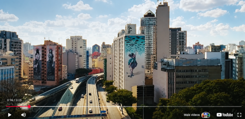Vista aérea do Centro de São Paulo com prédios altos, murais de arte urbana em grandes fachadas e o Elevado Presidente João Goulart (Minhocão) em primeiro plano. A imagem destaca a combinação entre arquitetura, mobilidade e intervenções artísticas que marcam a paisagem da região central.