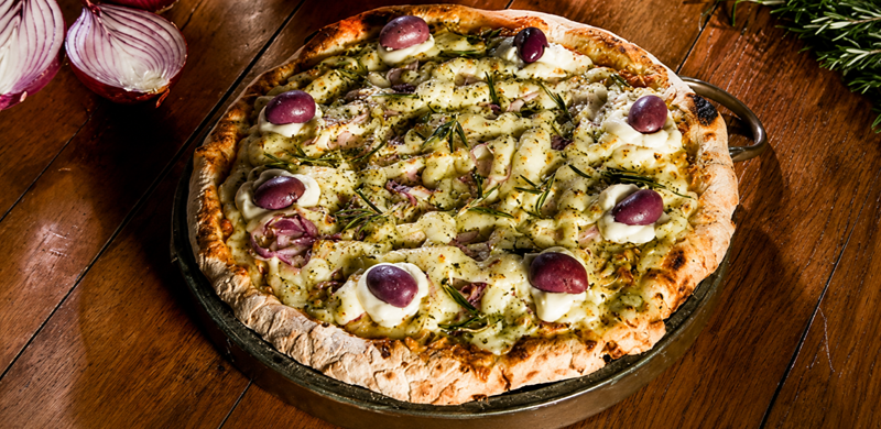 Pizza de borda alta servida em uma assadeira redonda sobre uma mesa de madeira. A cobertura é composta por queijo derretido, cebola-roxa, azeitonas-roxas inteiras, ervas frescas e temperos distribuídos por toda a superfície. No canto superior esquerdo, aparecem duas metades de cebola-roxa, enquanto ramos de alecrim decoram o canto superior direito, compondo um cenário rústico e acolhedor.