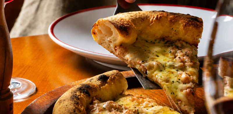 Fatia de pizza napolitana é erguida por uma espátula metálica sobre uma mesa de madeira. A borda alta e aerada revela a massa leve e bem fermentada, enquanto o queijo derretido forma fios entre a fatia e a pizza restante. Ao fundo, um prato branco com borda vermelha aparece desfocado, acompanhado por um moedor de madeira e uma taça, compondo um ambiente acolhedor de restaurante.