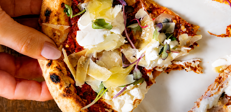 Close em uma fatia de pizza napolitana sendo segurada pela borda com a mão. A massa tem borda alta e levemente tostada, enquanto a cobertura reúne molho de tomate, burrata cremosa, lascas de queijo, folhas verdes e brotos. A fatia está sobre um prato branco, com parte da pizza ao fundo, destacando as texturas e os ingredientes frescos.