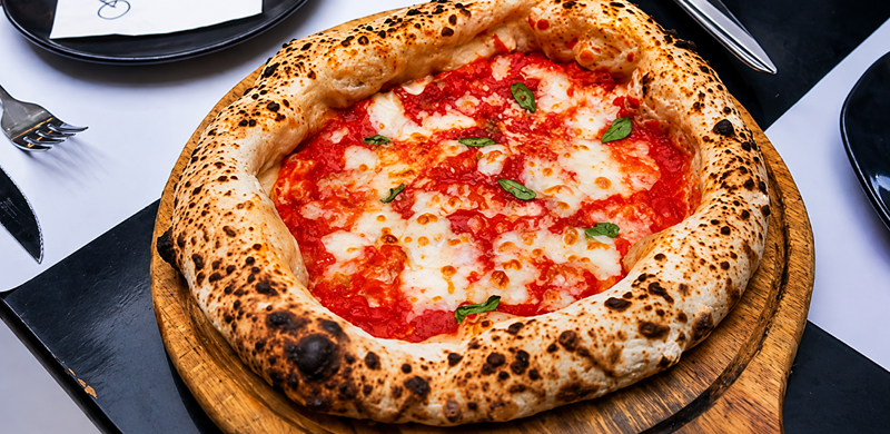 Pizza napolitana de muçarela e molho de tomate é servida sobre uma tábua de madeira. A borda alta e aerada apresenta marcas do forno a lenha, enquanto folhas de manjericão fresco decoram a cobertura. Ao redor, pratos, talheres e guardanapos compõem a mesa, destacando a pizza como protagonista da cena.