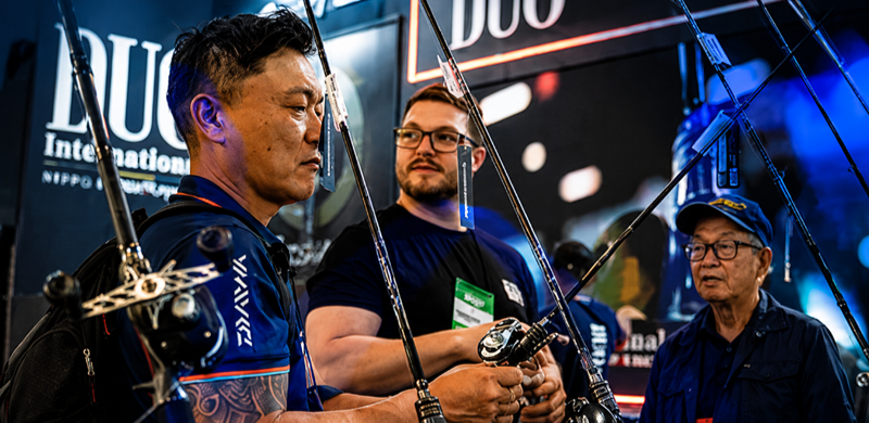 Visitantes observam e testam equipamentos de pesca durante a Fishing Show Brazil. Em primeiro plano, um expositor segura uma vara com carretilha enquanto conversa com outros participantes. Ao fundo, estandes de marcas do setor e iluminação em tons de azul reforçam o ambiente da feira.