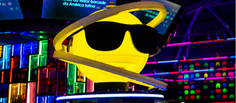 Ambiente interno iluminado por luzes em tons de azul, roxo, rosa e amarelo. Em destaque, uma escultura de emoji amarelo usando óculos escuros, cercada por um aro luminoso. Ao fundo, há painéis de LED, letreiros coloridos e áreas de jogos eletrônicos, criando uma atmosfera vibrante e futurista típica de um bar com fliperamas.