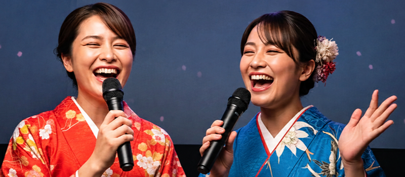 Duas mulheres sorriem enquanto cantam ao microfone em um palco. Uma veste um quimono vermelho com estampas florais e a outra usa um quimono azul com detalhes em branco e dourado, além de flores no cabelo. Ao fundo, há um painel em tom azul-escuro, destacando a cena de apresentação em um ambiente de karaokê.