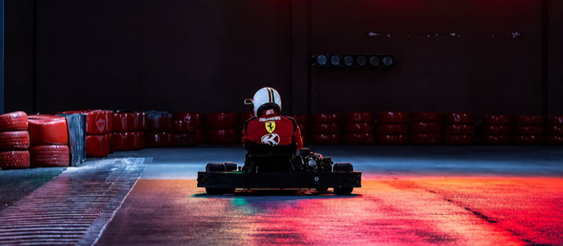 Piloto conduz um kart em uma pista coberta, visto de costas. O ambiente é iluminado por luzes vermelhas e azuis que se refletem no piso. Ao fundo, um grande painel eletrônico exibe posições e tempos da corrida, enquanto barreiras de pneus vermelhos delimitam a pista, criando um cenário que remete a uma competição de kart indoor.
