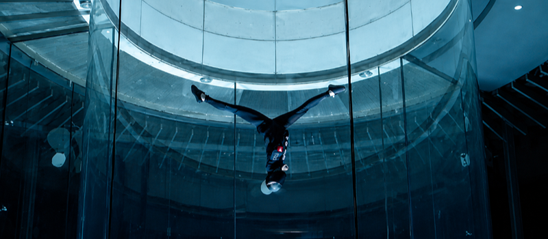Pessoa realiza voo indoor em um túnel de vento vertical de vidro na iFLY São Paulo. A imagem mostra o praticante de cabeça para baixo, com braços e pernas estendidos, no centro da estrutura circular iluminada por tons azulados. O ambiente moderno destaca a sensação de gravidade zero e a experiência de paraquedismo indoor.