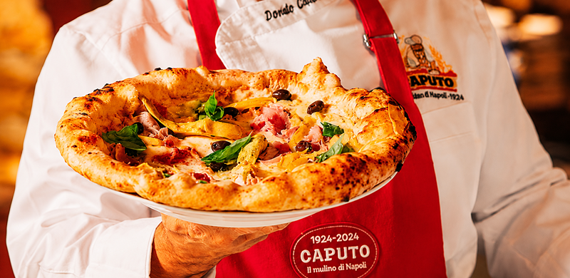 Pessoa vestindo dólmã branco e avental vermelho segura uma pizza napolitana sobre um prato branco. A pizza tem borda alta e dourada, levemente tostada, com cobertura de queijo derretido, presunto, azeitonas pretas, folhas de manjericão e outros ingredientes. O fundo desfocado, em tons quentes, remete ao ambiente interno de uma pizzaria, destacando a pizza como elemento principal da cena.
