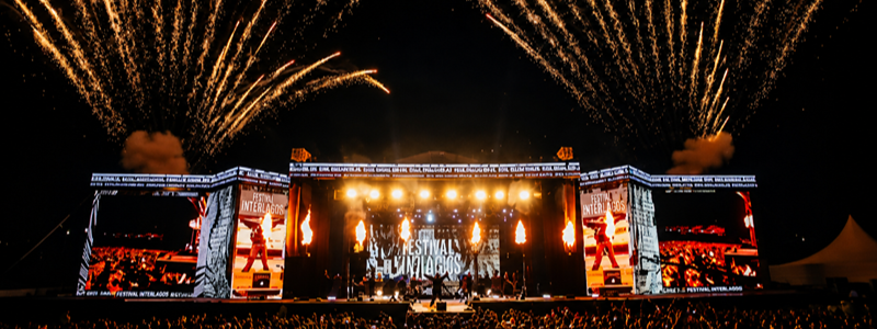 Imagem noturna de um grande palco montado ao ar livre durante o Festival Interlagos. Em primeiro plano, uma multidão acompanha o espetáculo de frente para o palco, iluminado por refletores em tons de amarelo e laranja. Nas laterais, dois telões exibem imagens da apresentação ao vivo. Acima da estrutura, fogos de artifício dourados cruzam o céu escuro em diferentes direções, formando um arco sobre o palco. Jatos de fogo também são lançados durante o show, reforçando o clima de celebração. A composição destaca a grandiosidade do evento, com milhares de pessoas reunidas para assistir às apresentações.