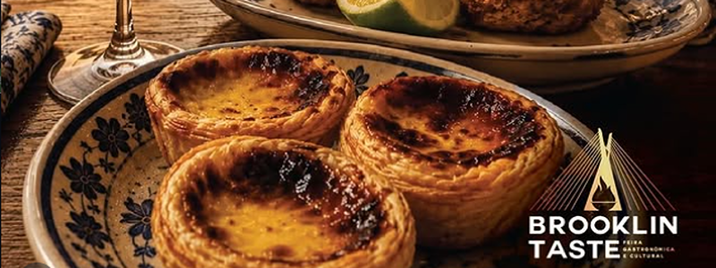 Imagem de uma mesa com pratos inspirados na gastronomia portuguesa. Em primeiro plano, três pastéis de nata estão servidos em um prato decorado em azul e branco. Ao lado, duas porções de bolinho de bacalhau acompanham uma fatia de limão e um ramo de ervas. Uma taça de vinho tinto completa a composição. Ao fundo, uma rua de paralelepípedos reúne restaurantes com mesas ao ar livre, bandeiras de Portugal e fachadas iluminadas por luzes amareladas, criando uma atmosfera acolhedora. Placas destacam um restaurante de culinária portuguesa e um quadro com sugestões de pratos típicos. No canto inferior direito, aparece o logotipo do Brooklin Taste.