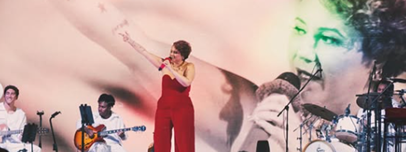 Maria Rita se apresenta no palco usando um macacão vermelho e segura um microfone enquanto canta com um dos braços erguidos. Ao fundo, um telão exibe um grande retrato da cantora em tons claros. A banda a acompanha com guitarra, bateria e outros instrumentos, diante de uma plateia parcialmente visível na parte inferior da imagem.
