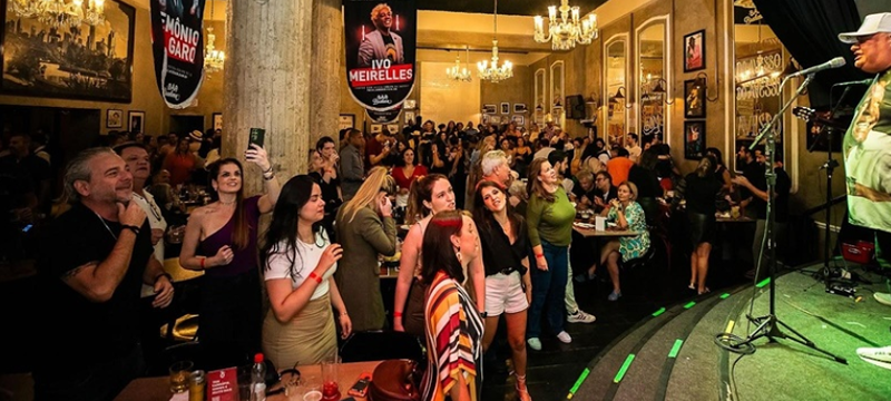 Público acompanha uma apresentação musical no Bar Brahma Centro. Pessoas ocupam mesas e ficam em pé próximas ao palco, onde um artista canta ao microfone. O salão tem lustres, quadros e cartazes de apresentações musicais nas paredes.